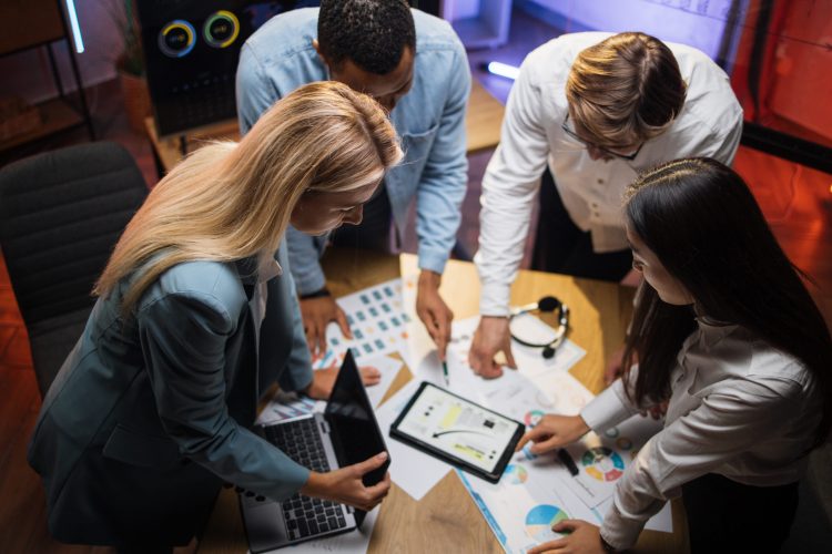 Top view of multiracial business partners analysing graphs and charts during briefing at modern office. Young people using modern tablets and laptops during conference.