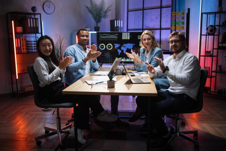 Multi ethnic male and female partners in formal clothes applauding together while sitting at office. Competent financiers with modern gadgets on table looking and smiling on camera.