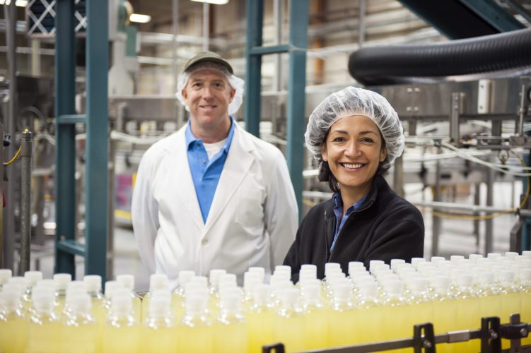 Portrait of a Caucasian male and African American female team of workers wearing head nets and standing near a conveyor belt of lemon flavored water in a bottling plant.